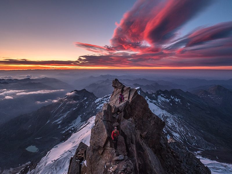 Climbers on a rocky summit ridge under red sunrise clouds